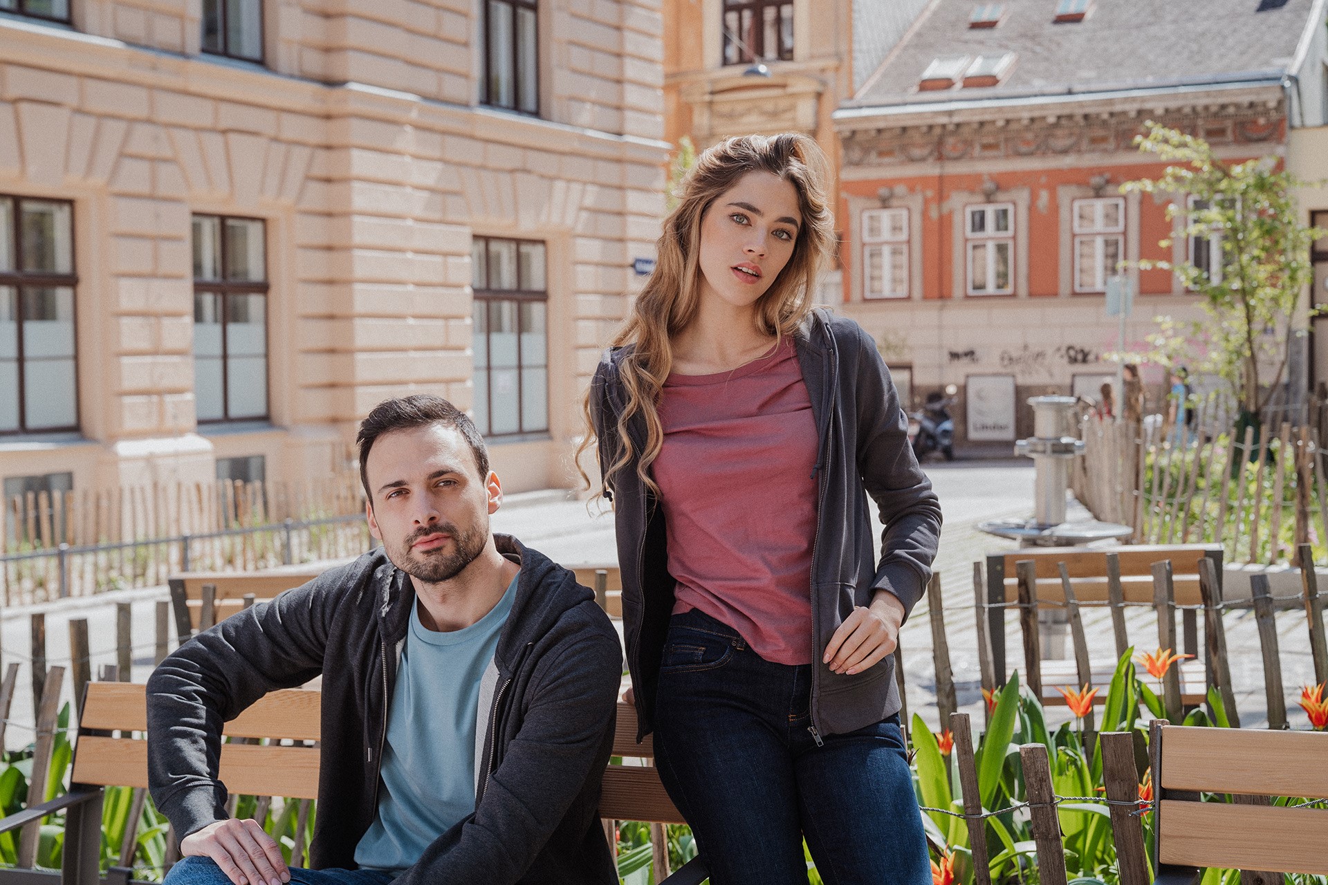 Junger Mann und junge Frau in nachhaltigen Jeans, lässig gekleidet, sitzen in einem urbanen Umfeld auf einer Holzbank.